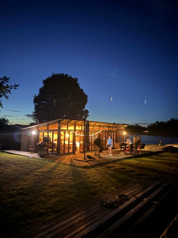 Stunning glass-walled pavilion glowing warmly at twilight, perfectly positioned by the water's edge. This magical retreat offers panoramic views and seamless indoor-outdoor living, ideal for evening gatherings under the starlit Swedish sky.